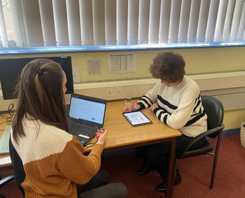 Two women sitting at a desk. One lady is timing the other while she fills out some information on an IPad.