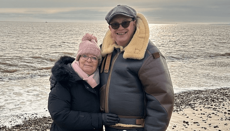 Two people standing on a beach hugging, looking at the camera smiling