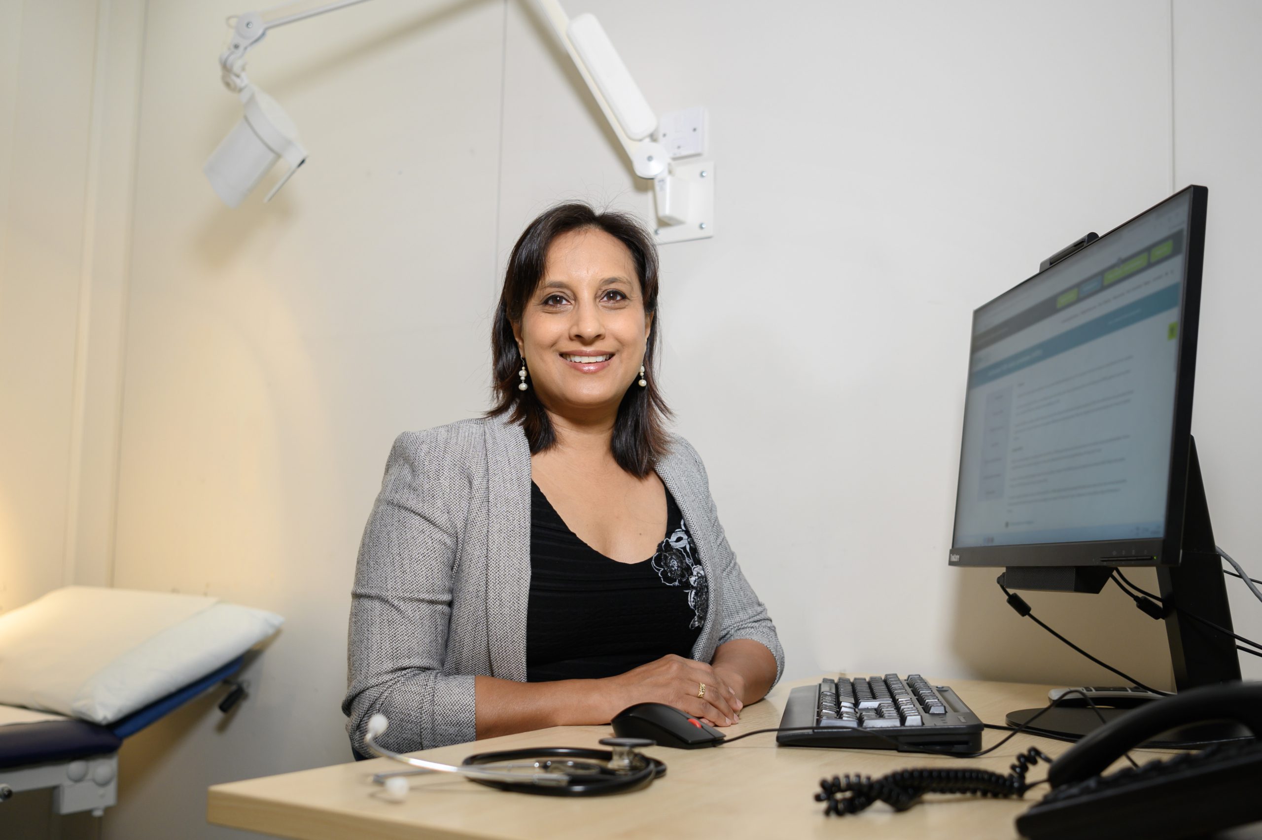 woman in a grey jacket and black top sitting at a desk, with a PC, turned towards the camera smiling