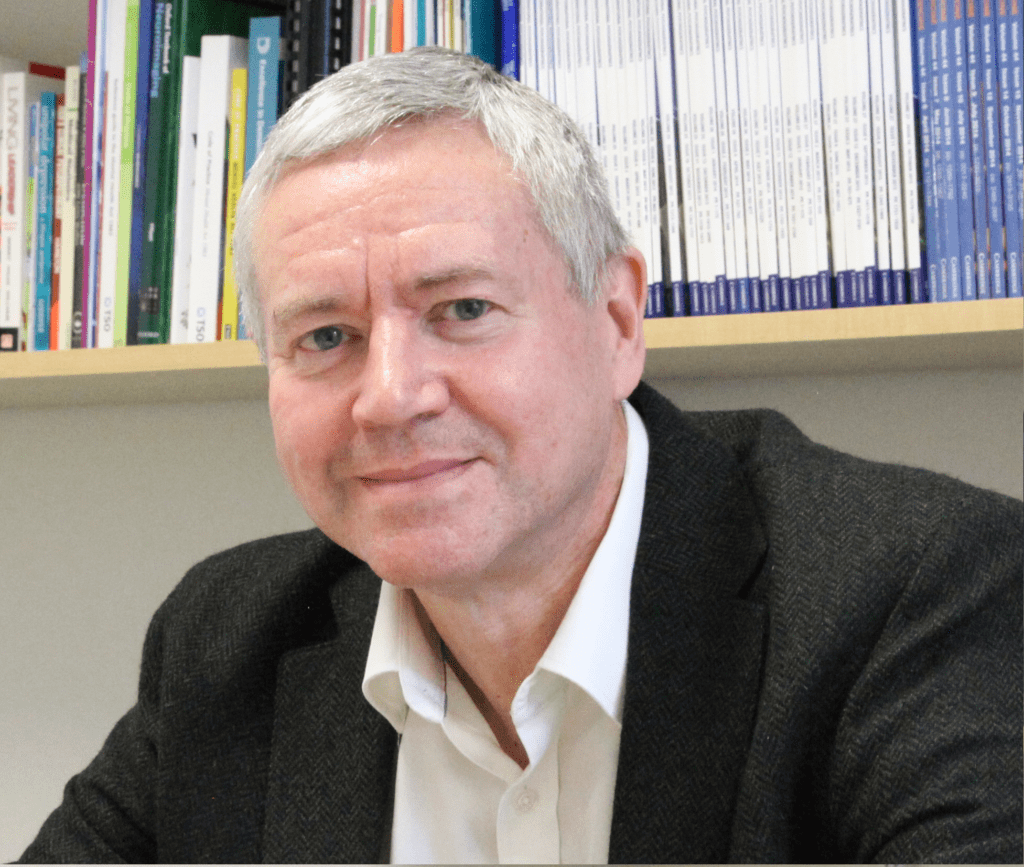 Man in a suit sitting in front of a book case looking at the camera smiling.