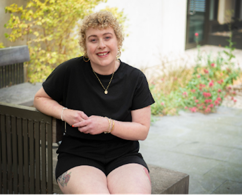 Woman in a black t-shirt and shorts, sitting on a bench, looking at the camera smiling